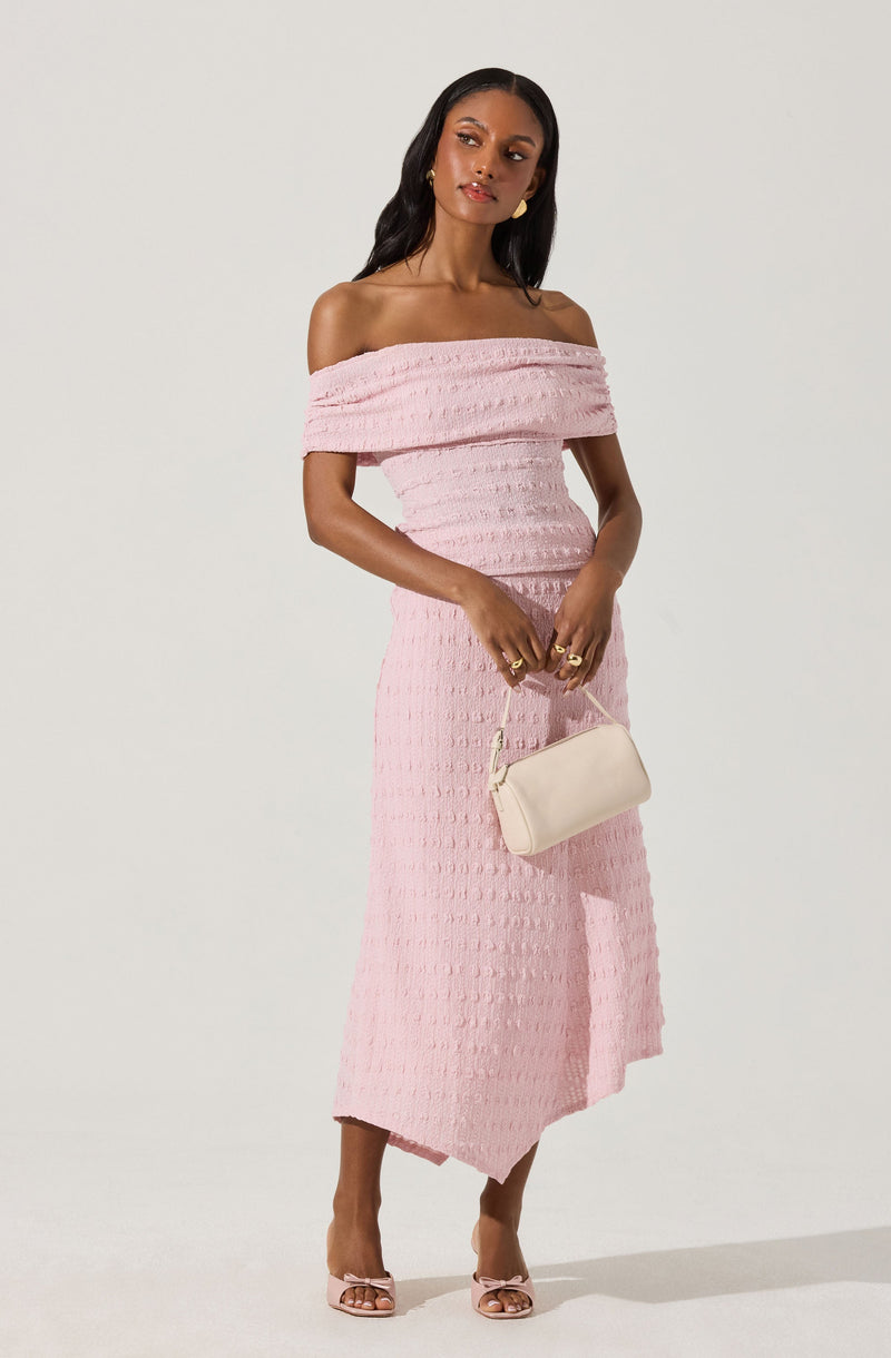 A woman is wearing a light pink, off-the-shoulder dress with a textured, ribbed pattern. She is holding a small, cream-colored handbag and is wearing open-toed, pink high-heeled sandals with bow details.
