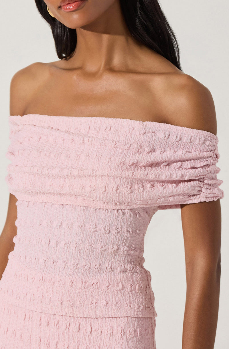 A woman is wearing a light pink, off-the-shoulder dress that features a textured, ruched fabric with horizontal bands and small, raised details throughout. The dress has a fitted silhouette and exposes her shoulders and collarbone.