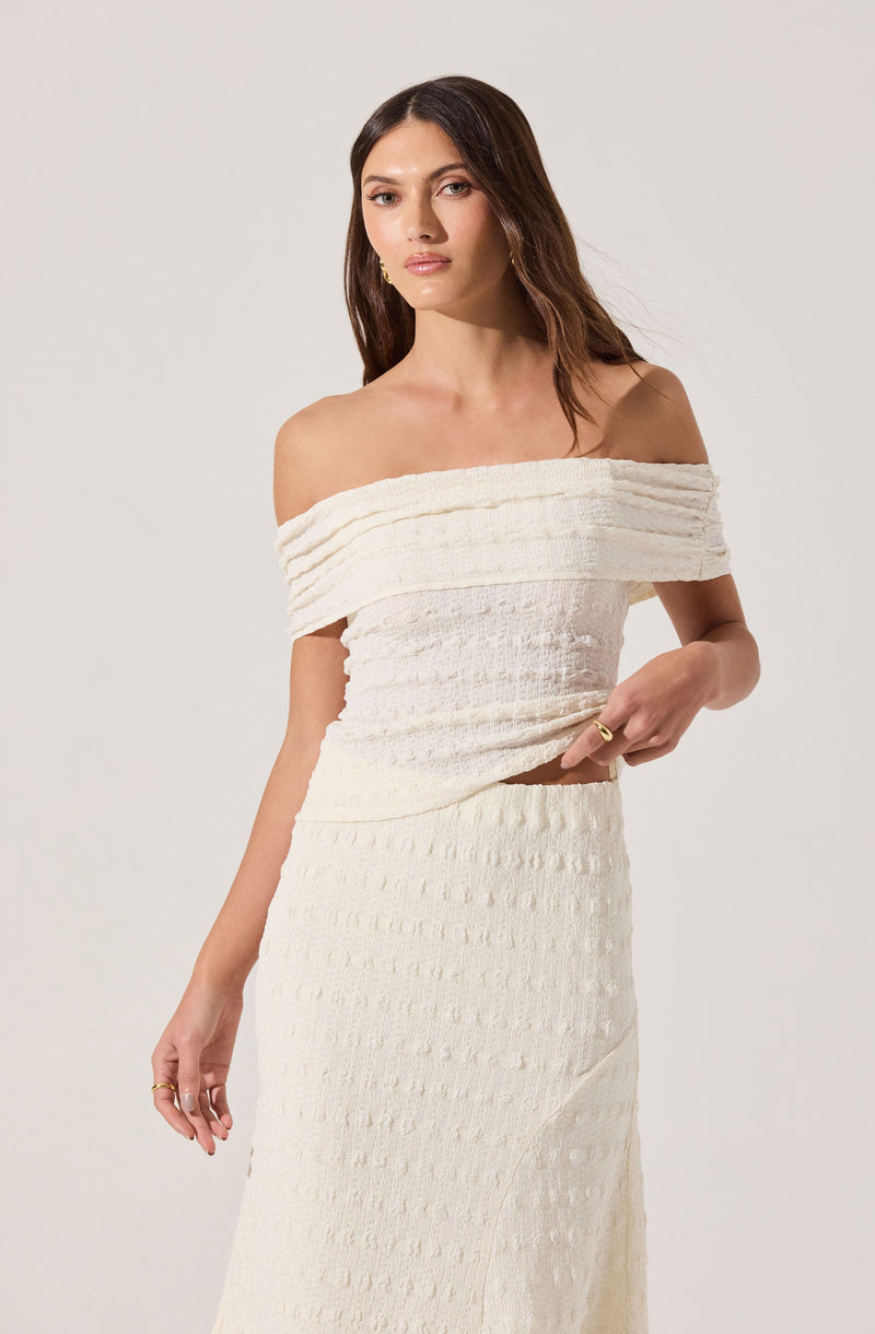 A woman is wearing an off-the-shoulder, cream-colored dress with textured horizontal stripes, featuring a fitted bodice and a long skirt.