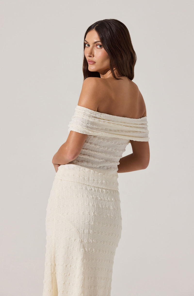 A woman wearing an off-the-shoulder, textured, cream-colored dress with short sleeves that drape over her upper arms. The dress has a fitted bodice and a long, flowing skirt with a subtle horizontal pattern.