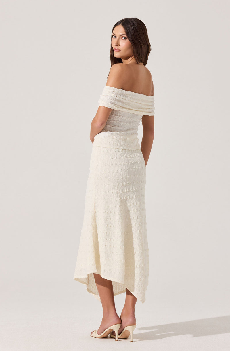 A woman is wearing an off-the-shoulder, cream-colored textured dress that is fitted at the top and flows into a longer skirt with a subtle pattern. She is also wearing nude high-heeled shoes.