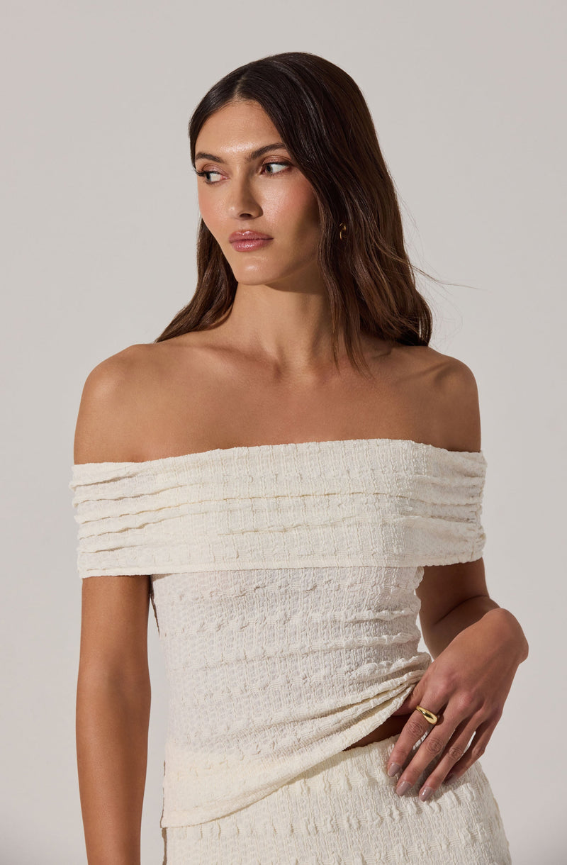 A woman is wearing an off-the-shoulder, cream-colored, textured knit top with horizontal ribbing. The top appears to be made of a soft, stretchy fabric that fits snugly around her torso. The texture of the fabric is slightly scrunched, adding visual interest to the garment.