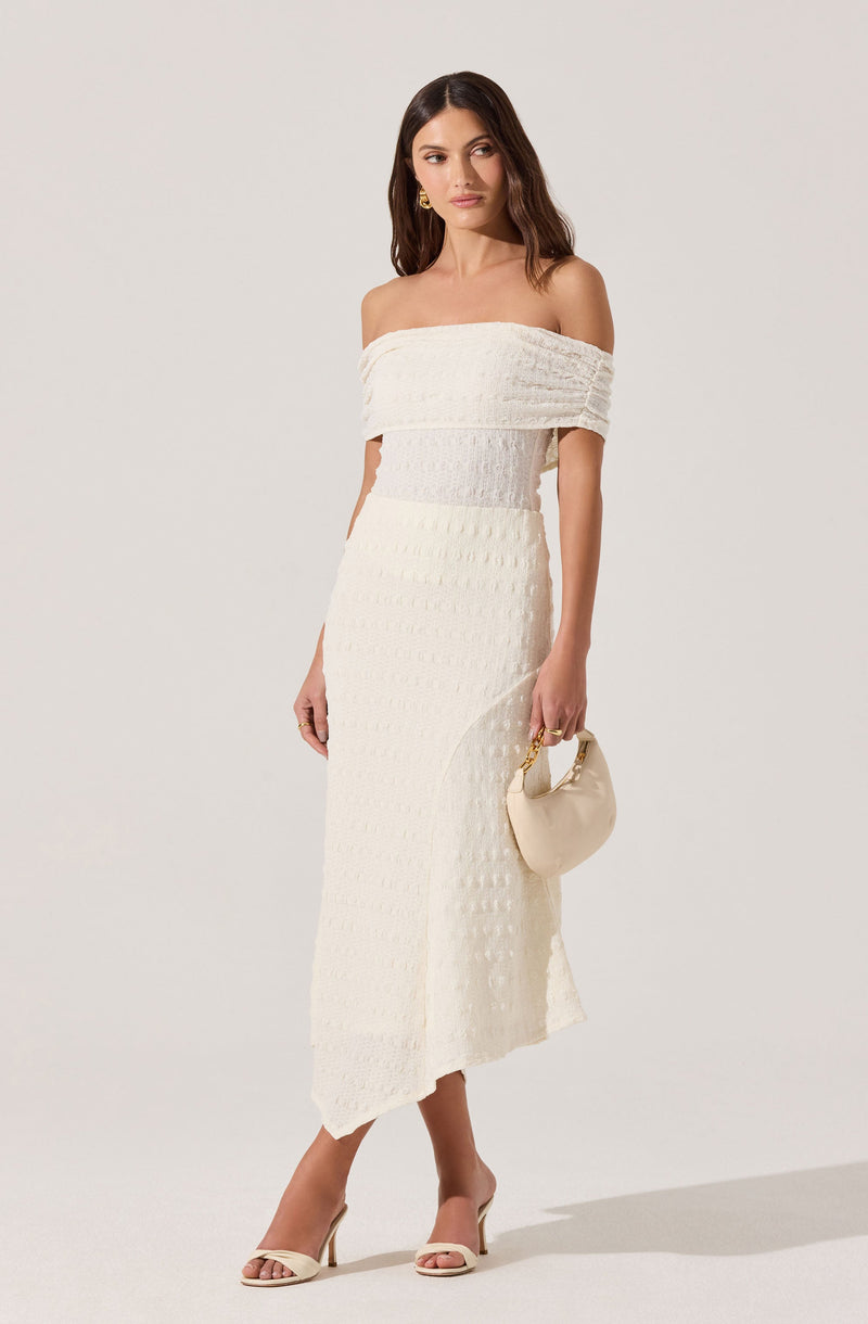 A woman is wearing an off-the-shoulder white textured dress that is fitted at the waist and extends to a mid-calf length with a slit on one side. She is holding a small, light-colored handbag with a gold handle, and is wearing beige high-heeled sandals. She has long, dark, wavy hair and is accessorizing with gold earrings.