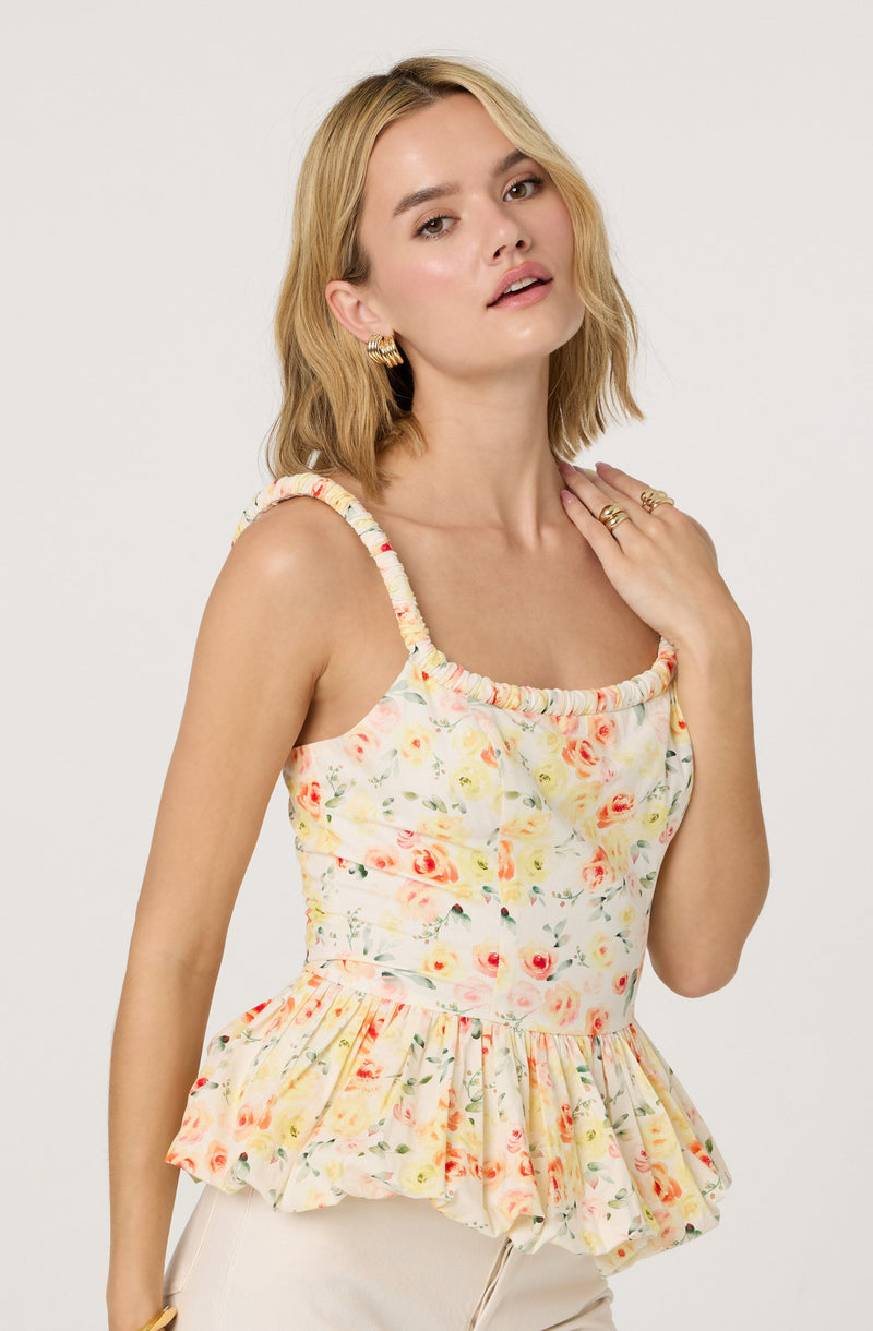 A woman is wearing a sleeveless, floral print top with ruffled straps and a ruffled hem. The top features a light background with watercolor-style roses in shades of pink, orange, and yellow, along with green leaves.