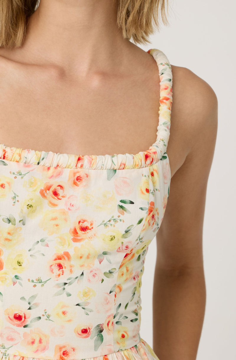 Close-up of a woman's upper body wearing a sleeveless, floral-printed dress with spaghetti straps. The dress features a gathered, elasticized neckline with a soft, ruffled edge. The fabric displays a pattern of pastel-colored flowers in shades of pink, yellow, orange, and green on a light background.