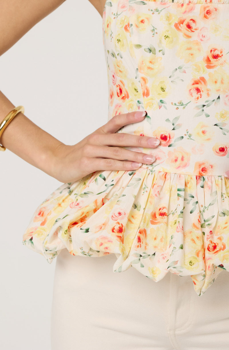 A woman is wearing a sleeveless, floral print top with a ruffled hem. The top features a pattern of pink, orange, and yellow flowers with green leaves on a light background. She has a gold bracelet on her left wrist and beige pants.