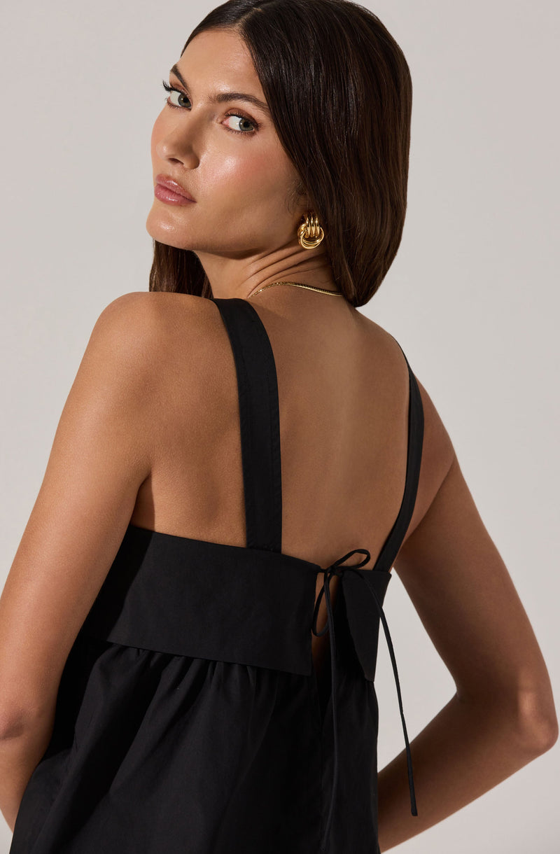 A woman is wearing a black sleeveless dress with wide straps and a large bow tied at the back. The dress has a gathered skirt and a structured bodice. She has gold hoop earrings and a delicate gold necklace.