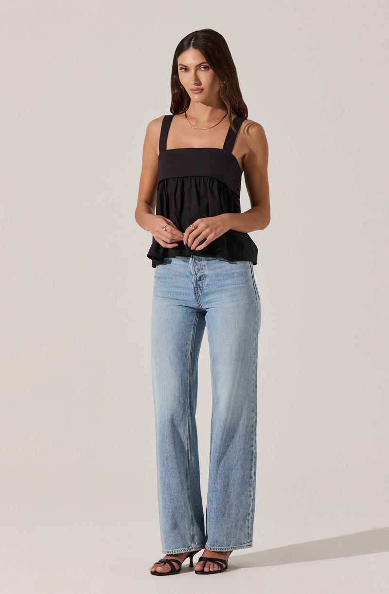 A woman is wearing a black sleeveless top with wide straps, featuring a gathered, ruffled design at the bottom. She pairs it with light blue, high-waisted, wide-leg jeans and black high-heeled sandals. She has long, dark hair and is posing with her hands gently clasped in front of her.