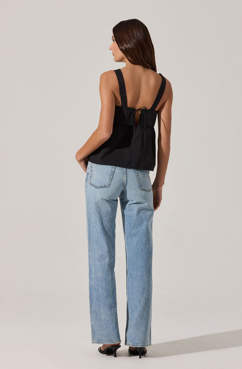 A woman is wearing a sleeveless black top with wide straps and a tied bow at the back, paired with light blue high-waisted, straight-leg jeans. She is also wearing black high-heeled shoes.