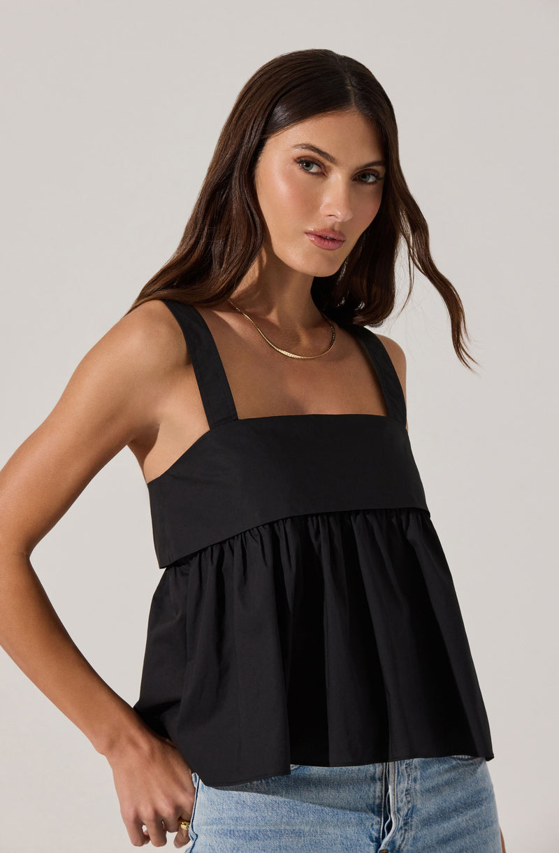 A woman is wearing a black sleeveless top with wide shoulder straps, featuring a sheer panel at the neckline and a gathered, loose-fitting design from the bust down.