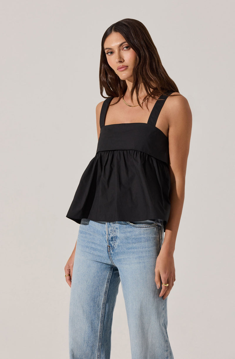 A woman is wearing a black sleeveless top with wide shoulder straps, featuring a square neckline and a gathered, flared hem. She pairs it with light-wash, high-waisted jeans.