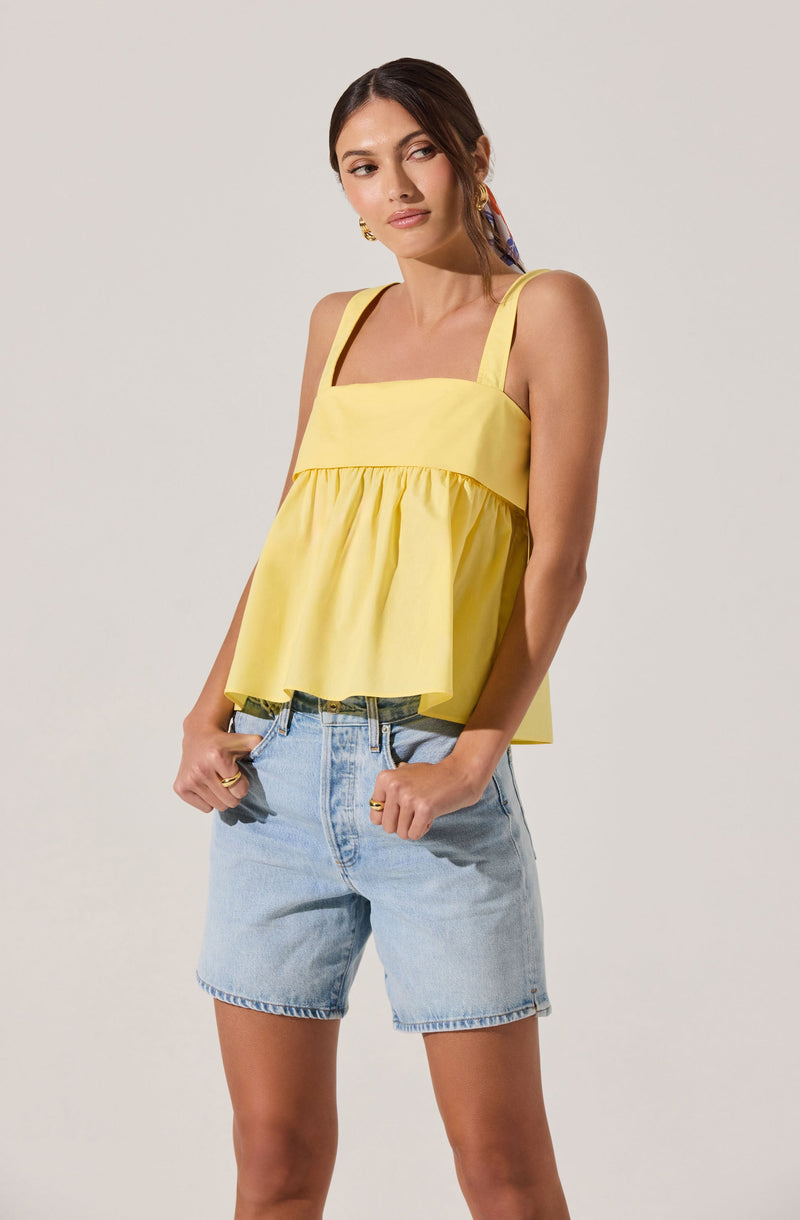A woman is wearing a sleeveless, bright yellow top with a square neckline and wide shoulder straps. The top has a loose, ruffled hem that creates a flowy, relaxed fit. She pairs it with light-wash denim shorts that have a classic five-pocket design and a frayed hem.