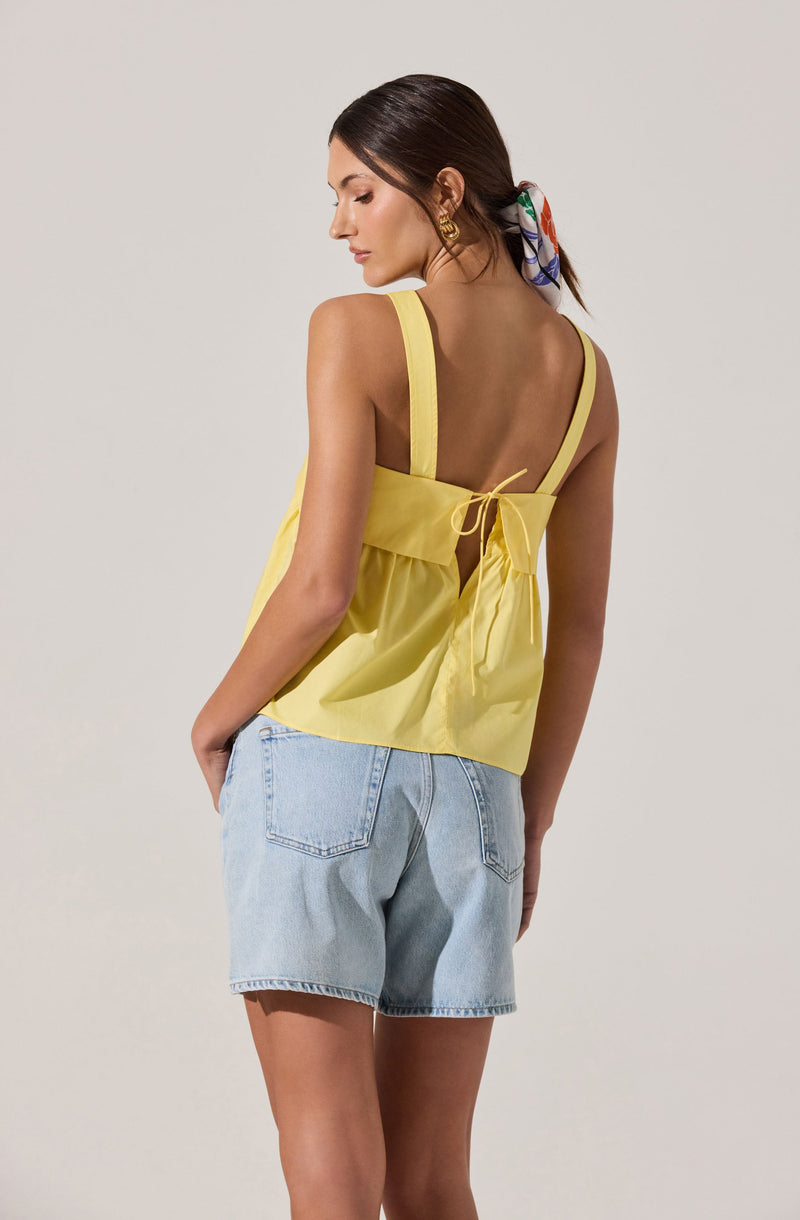 A woman is wearing a sleeveless yellow top with wide shoulder straps and a back tie detail, paired with light blue denim shorts. She has a colorful scarf in her hair, and is accessorized with gold earrings.