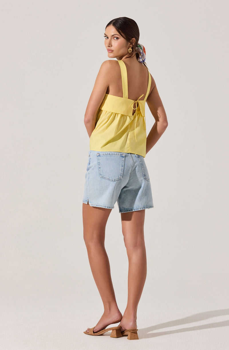 A woman is wearing a yellow sleeveless top with wide shoulder straps and a tied bow at the back. She has paired it with light-wash denim shorts and beige open-toe sandals with a block heel. She also has large hoop earrings and a colorful scarf tied in her hair.