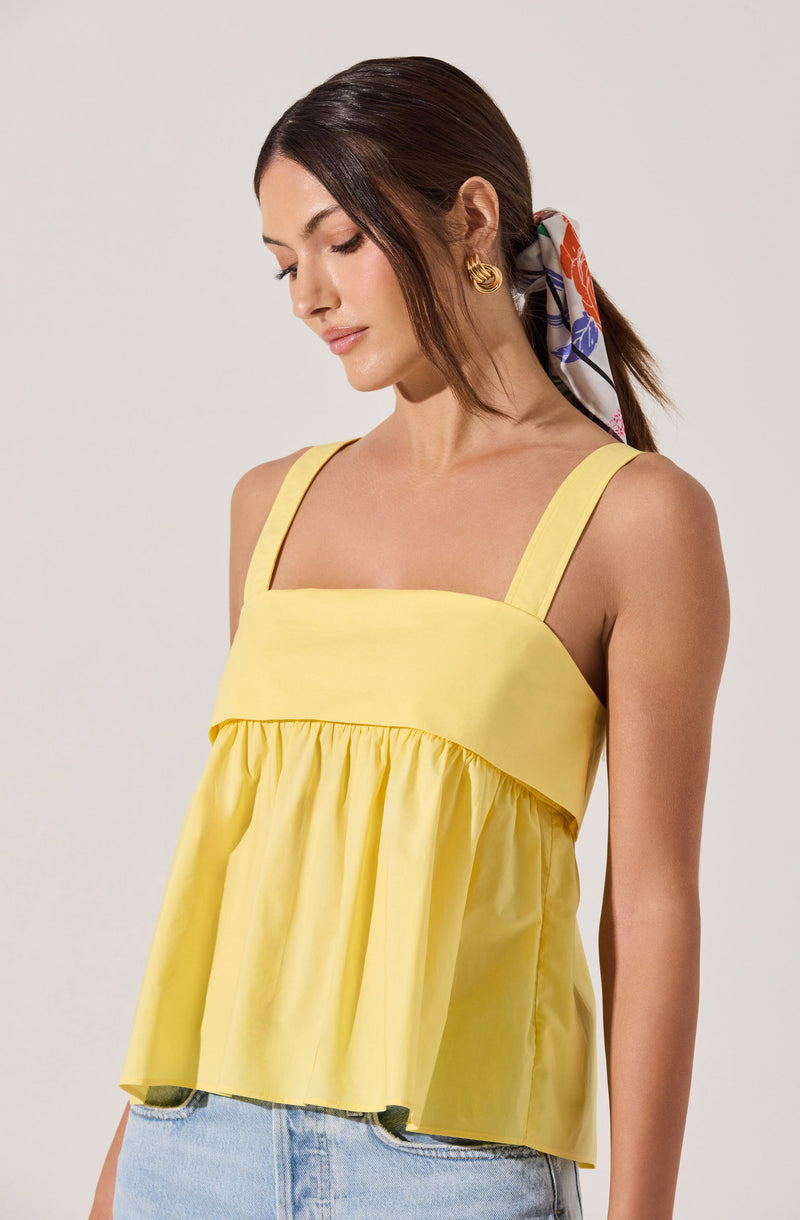 The woman is wearing a sleeveless yellow top with wide straps and a gathered, ruffled hem. She has paired it with light blue jeans. She accessorizes with gold hoop earrings and a colorful scarf tied in her hair.