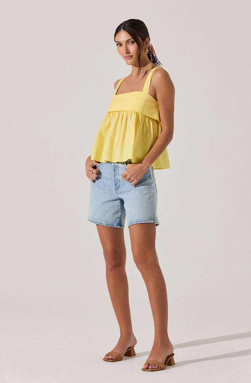 A woman is wearing a sleeveless yellow top with wide shoulder straps and a loose, flowy fit. She has paired it with light blue denim shorts that have a relaxed fit and a frayed hem. She is also wearing beige open-toe heeled sandals.