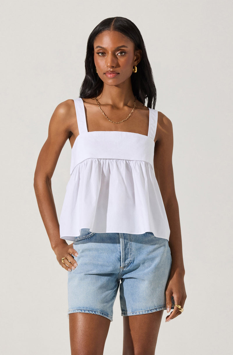 A woman is wearing a white sleeveless top with wide straps, featuring a fitted bodice and a gathered, flared lower section. She pairs it with light blue denim shorts.