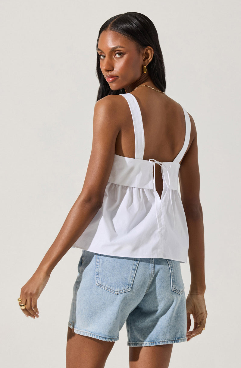 A woman is wearing a white sleeveless top with wide shoulder straps, a gathered waist detail, and a keyhole opening at the back tied with a small bow. She is also wearing light blue denim shorts with visible stitching and a slightly frayed hem. She has gold hoop earrings and a gold chain necklace.