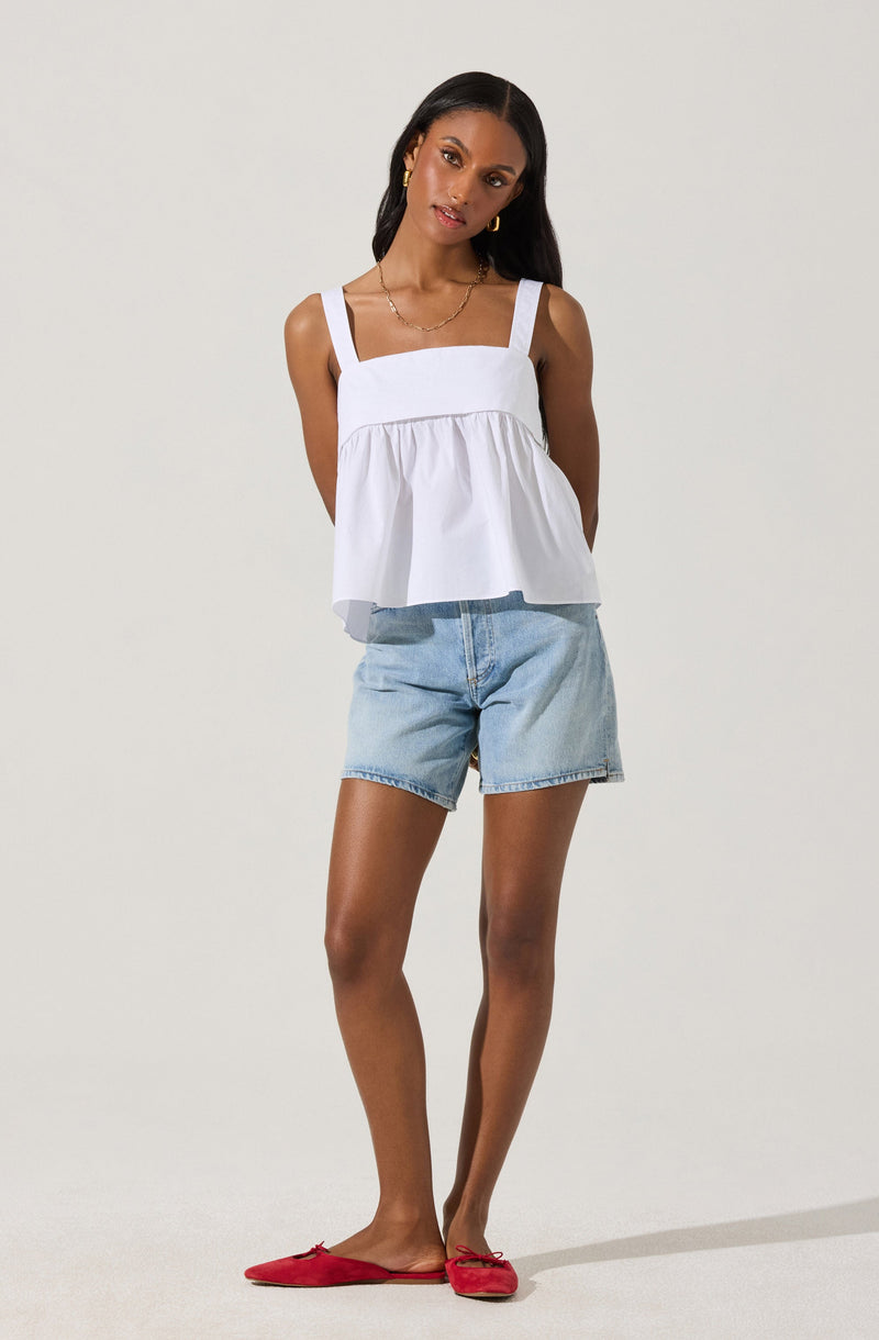 A woman is wearing a white sleeveless top with wide shoulder straps and a gathered, ruffled hem. She has on light blue denim shorts and red flat shoes with small bows on the toes.