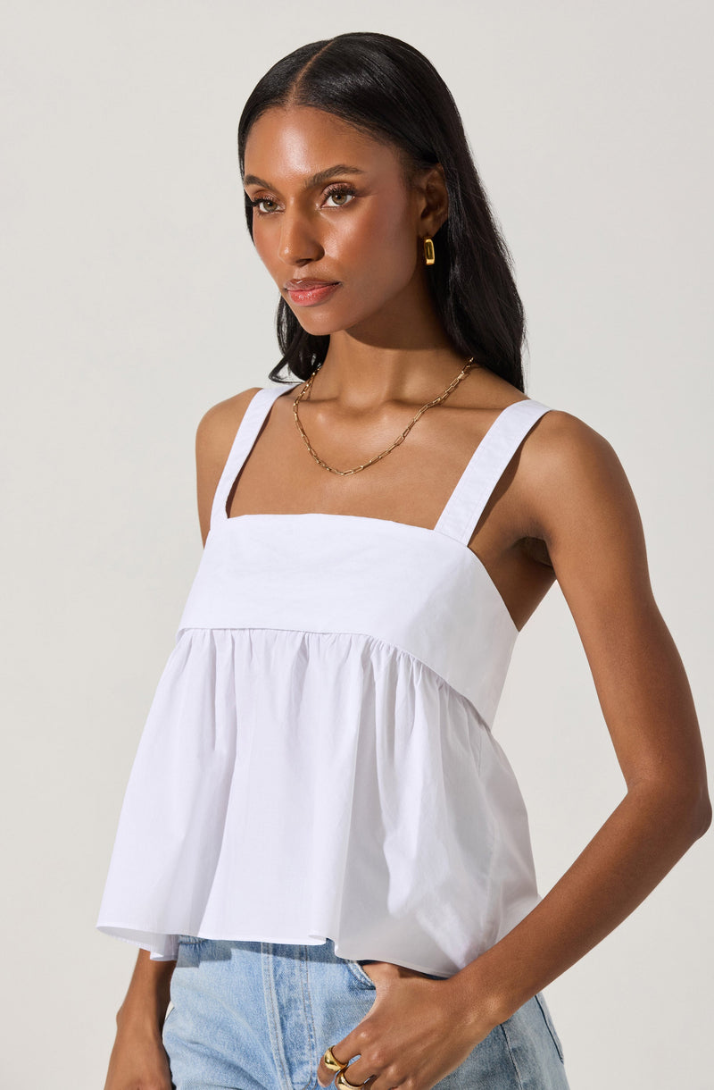 A woman is wearing a white sleeveless top with wide shoulder straps and a square neckline, featuring a gathered, loose-fitting design that extends below the bust. She is also wearing light blue denim jeans. The outfit is accessorized with gold jewelry, including hoop earrings, a chain necklace, and rings.