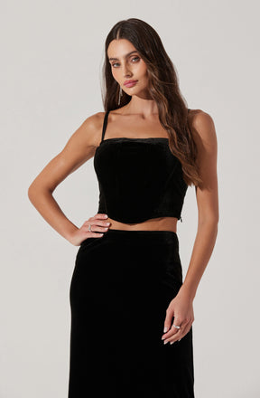 A woman is wearing a black velvet sleeveless crop top with thin straps and a high-waisted black velvet skirt.