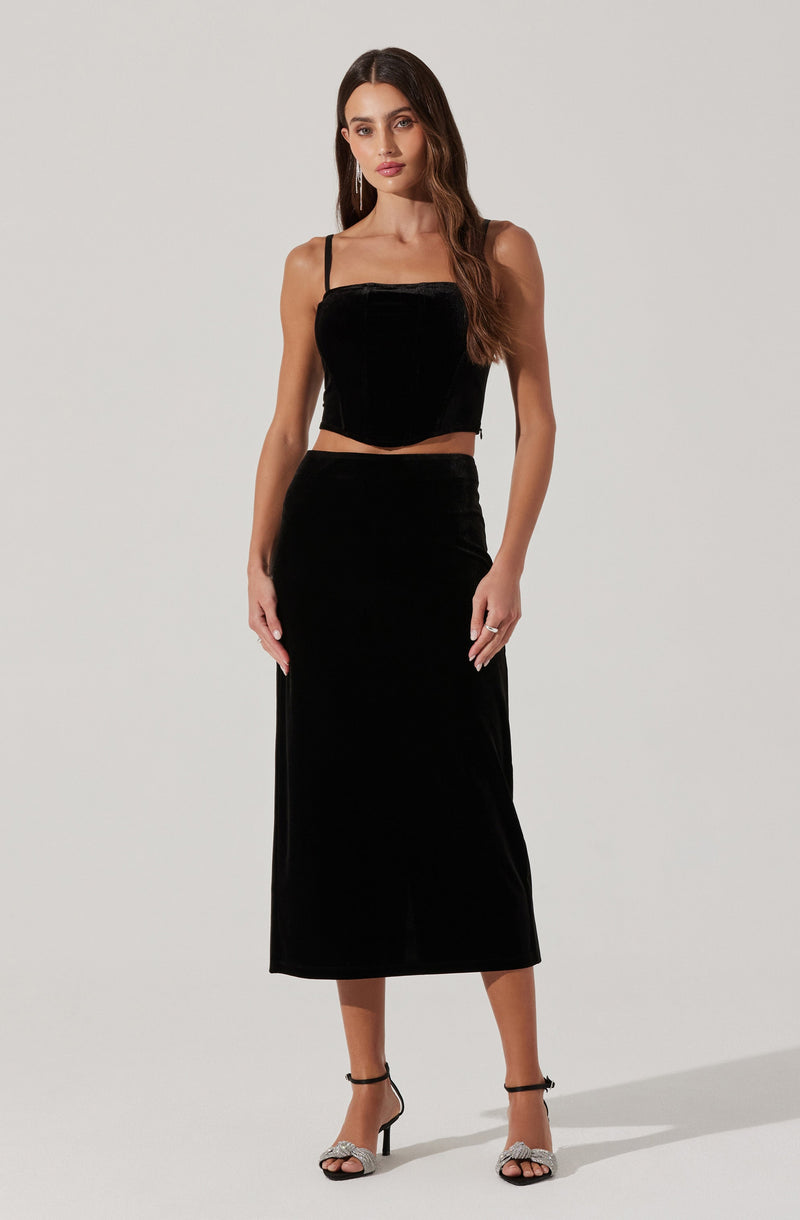 A woman is wearing a black outfit consisting of a sleeveless, square-neck crop top with thin spaghetti straps and a long, high-waisted velvet skirt that reaches her mid-calf. She is also wearing black high-heeled sandals with ankle straps, featuring a bow detail on the front.