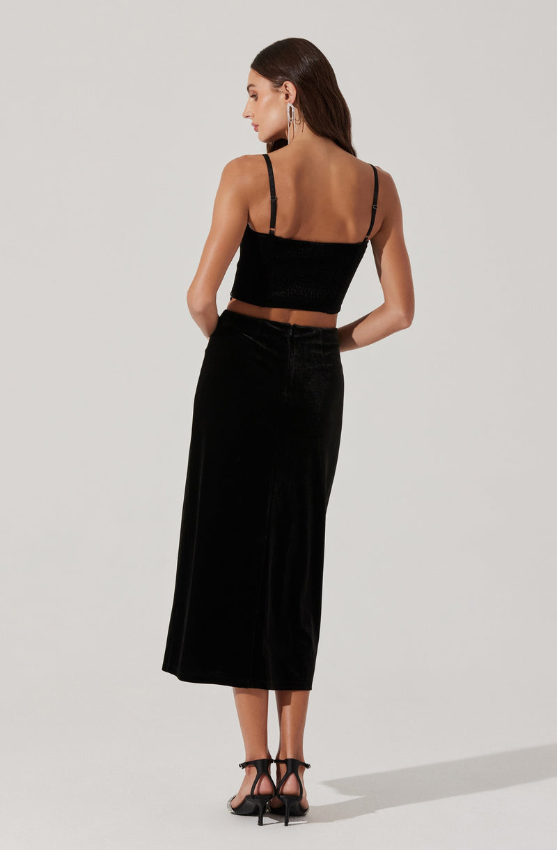 A woman is wearing a black spaghetti strap crop top with a textured fabric and matching high-waisted black velvet midi skirt with a straight cut. She is also wearing black high-heeled sandals with ankle straps.