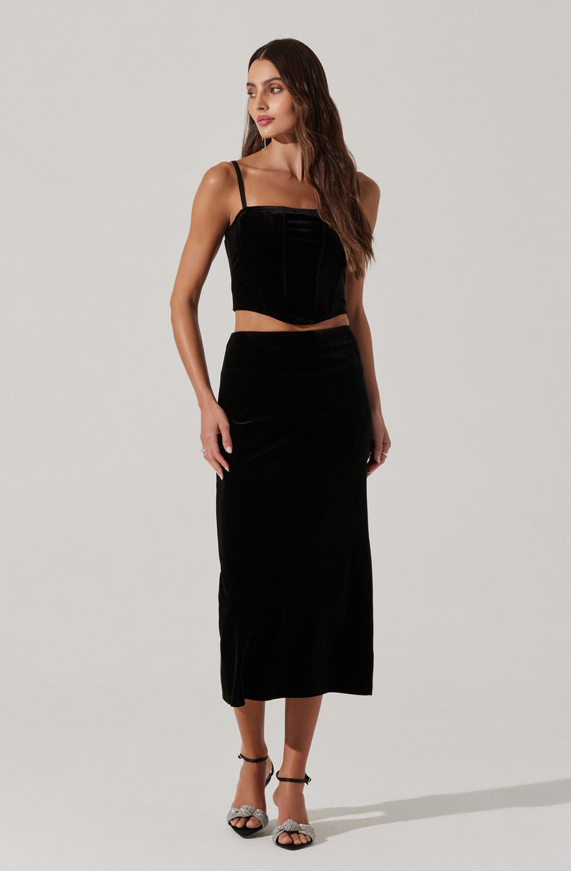A woman is wearing a black outfit consisting of a sleeveless, fitted crop top with thin spaghetti straps and a high-waisted, ankle-length skirt made of a shiny, velvet-like material. She is also wearing black high-heeled sandals with ankle straps that have decorative bows.