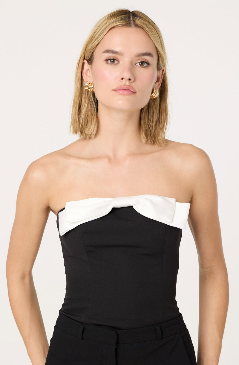 A woman is wearing a strapless black top with a large white bow across the chest. She is accessorized with gold hoop earrings.