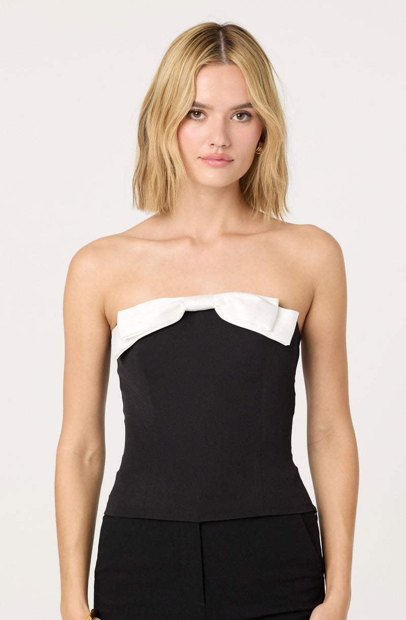 A woman is wearing a strapless black top with a large white bow detail across the top edge. The top has a fitted silhouette, and she pairs it with matching black bottoms.