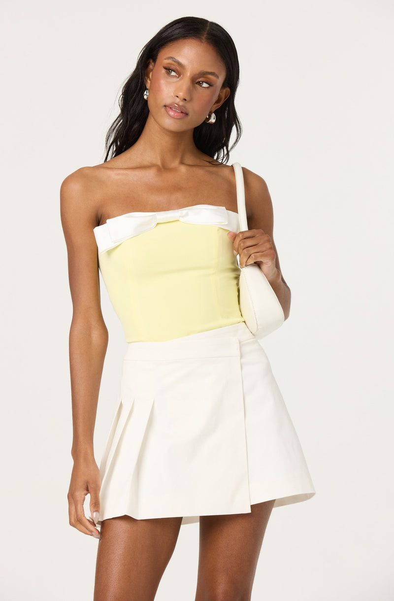 A woman is wearing a sleeveless, strapless, light yellow top with a large white bow detail across the top edge. She pairs it with a high-waisted, white A-line skirt that features pleats on one side. She is carrying a small white shoulder bag and is wearing pearl earrings.