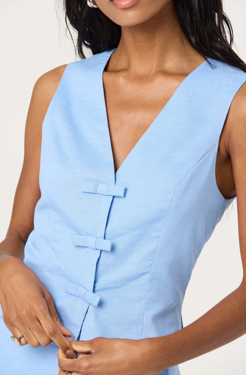 A woman is wearing a sleeveless, light blue dress with a V-neckline. The dress features traditional Chinese frog closures down the front. The material appears to be a smooth, woven fabric.