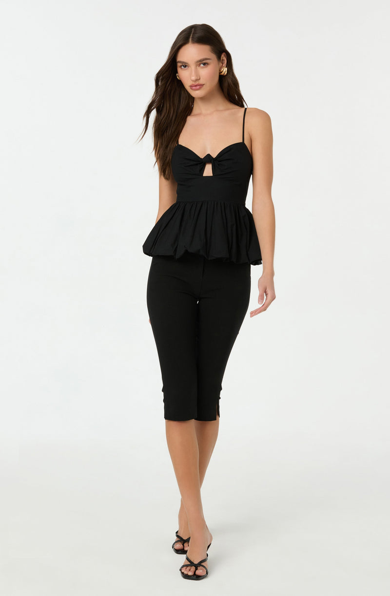 A woman is wearing a black sleeveless top with spaghetti straps, featuring a bow and a keyhole cutout at the front, and a ruffled peplum waist. She pairs it with black cropped pants and black high-heeled sandals.
