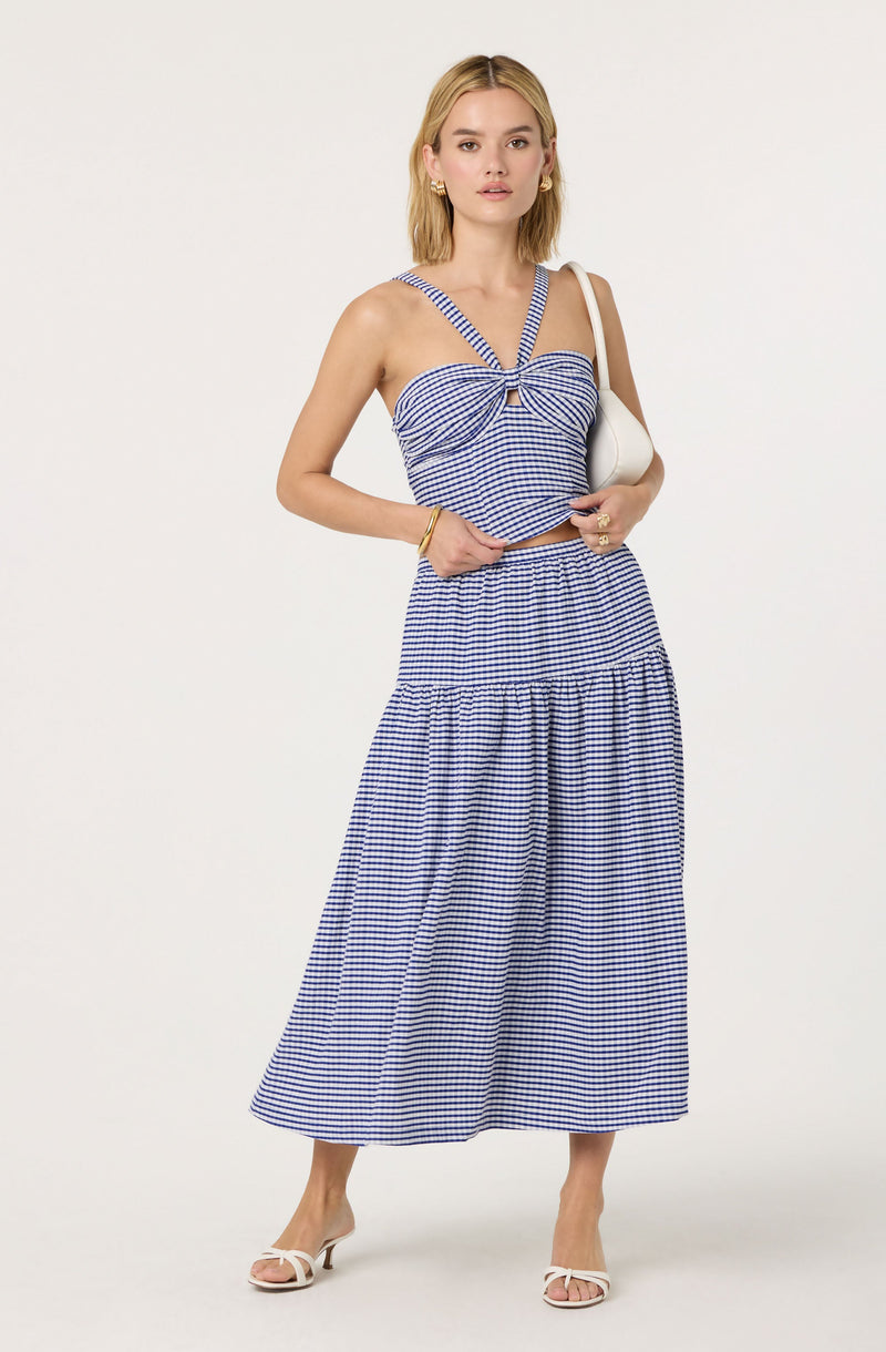 A woman is wearing a blue and white checkered two-piece outfit, consisting of a sleeveless top with shoulder straps and a bow detail at the front, and a matching long, wide-hem skirt with a gathered waist. She pairs the outfit with white heeled sandals and accessories including gold hoop earrings, a bracelet, and rings, and carries a small white shoulder bag.
