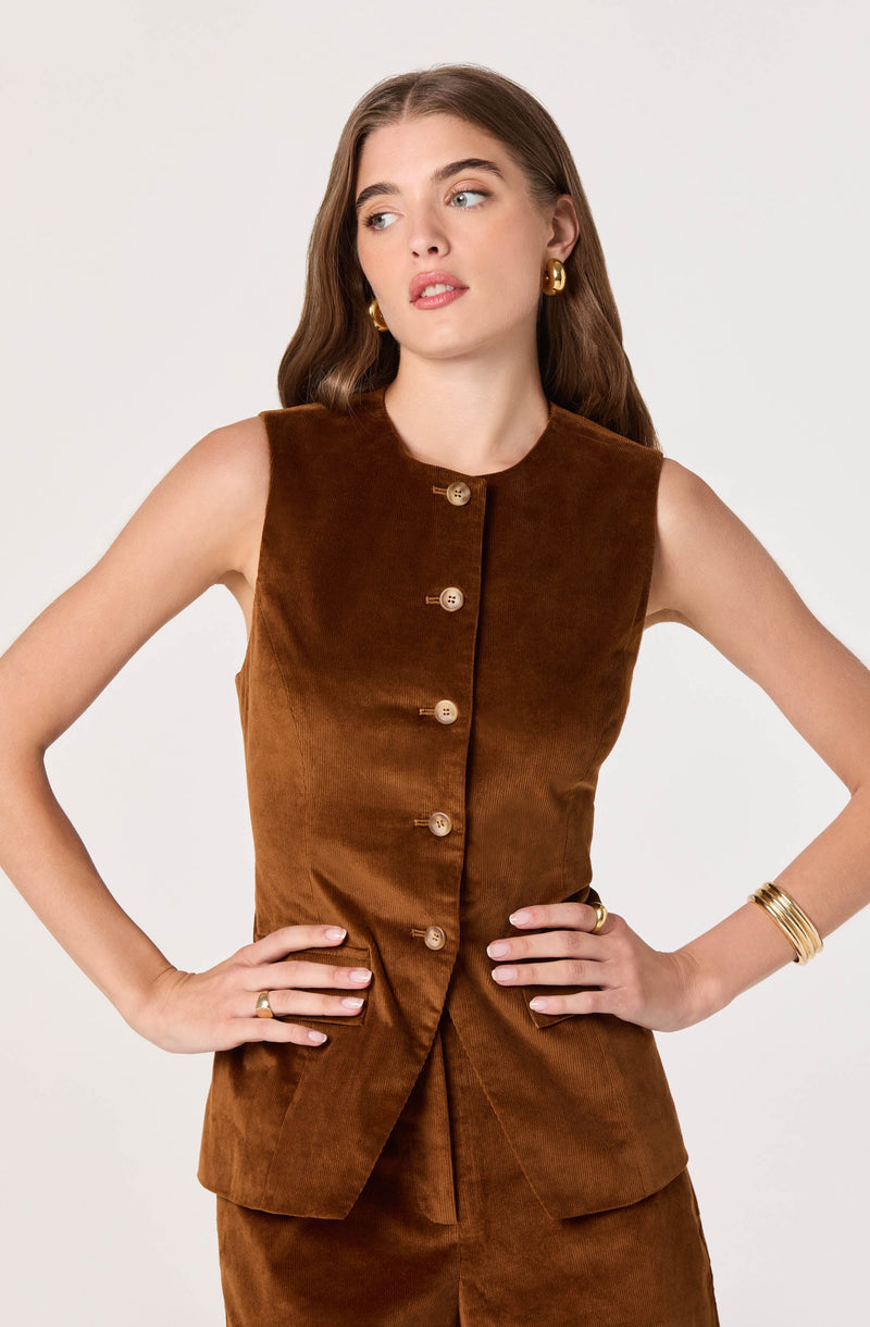 A woman is wearing a sleeveless, brown velvet vest with a round neckline and six light-colored buttons down the front. She pairs it with matching brown velvet pants. She has gold hoop earrings, a gold ring on her left hand, and multiple gold bangles on her right wrist.