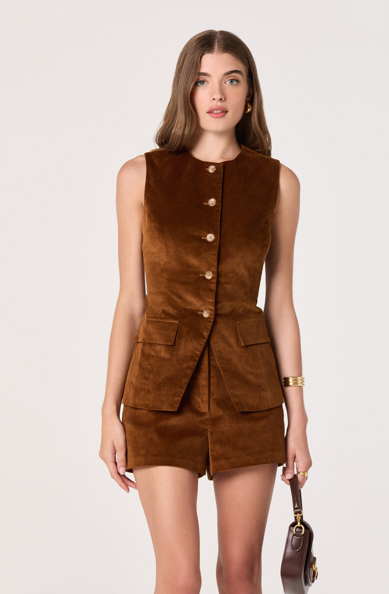A woman is wearing a sleeveless, brown suede vest with a round neckline and gold buttons down the front. The vest has two flap pockets on the front at hip level. She is also wearing a matching brown suede mini skirt. She accessorizes with gold earrings, a gold bracelet, and holds a small brown handbag with a gold chain strap.