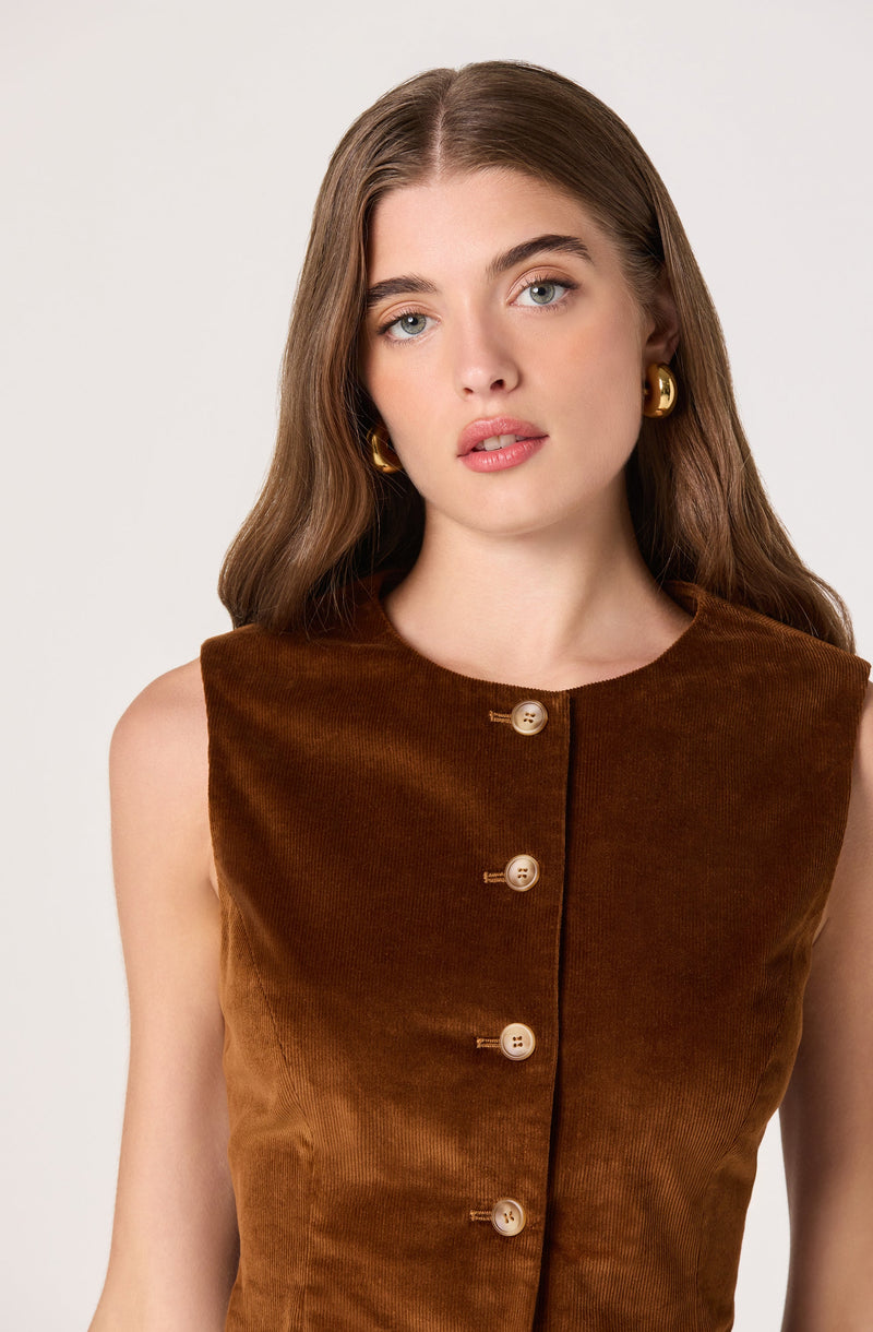 A woman with long wavy brown hair is wearing a sleeveless, brown velvet top with a round neckline. The top features a row of large, round, beige buttons down the front. She has gold hoop earrings and natural makeup.