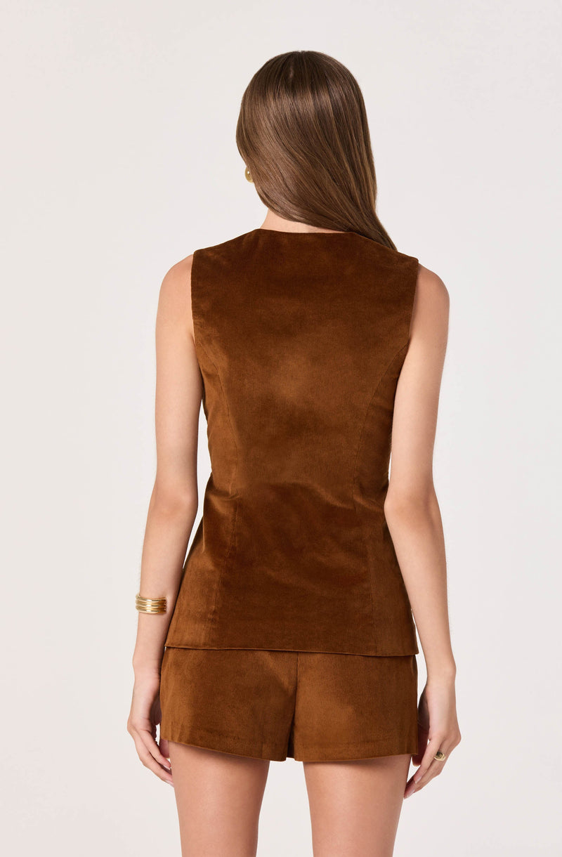 The image shows a woman wearing a sleeveless, brown suede top and matching shorts. The top has a high neckline and a fitted silhouette. The shorts are also made of suede and have a tailored fit. She is accessorized with a gold bracelet on her right wrist and a ring on her right hand. The background is plain and white.