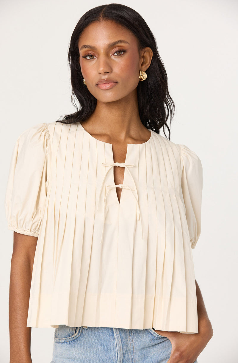 A woman is wearing a cream-colored, pleated blouse with short puff sleeves. The blouse features vertical pleats and a round neckline with a small keyhole opening fastened by two ties. She is also wearing light blue denim jeans and has gold hoop earrings.