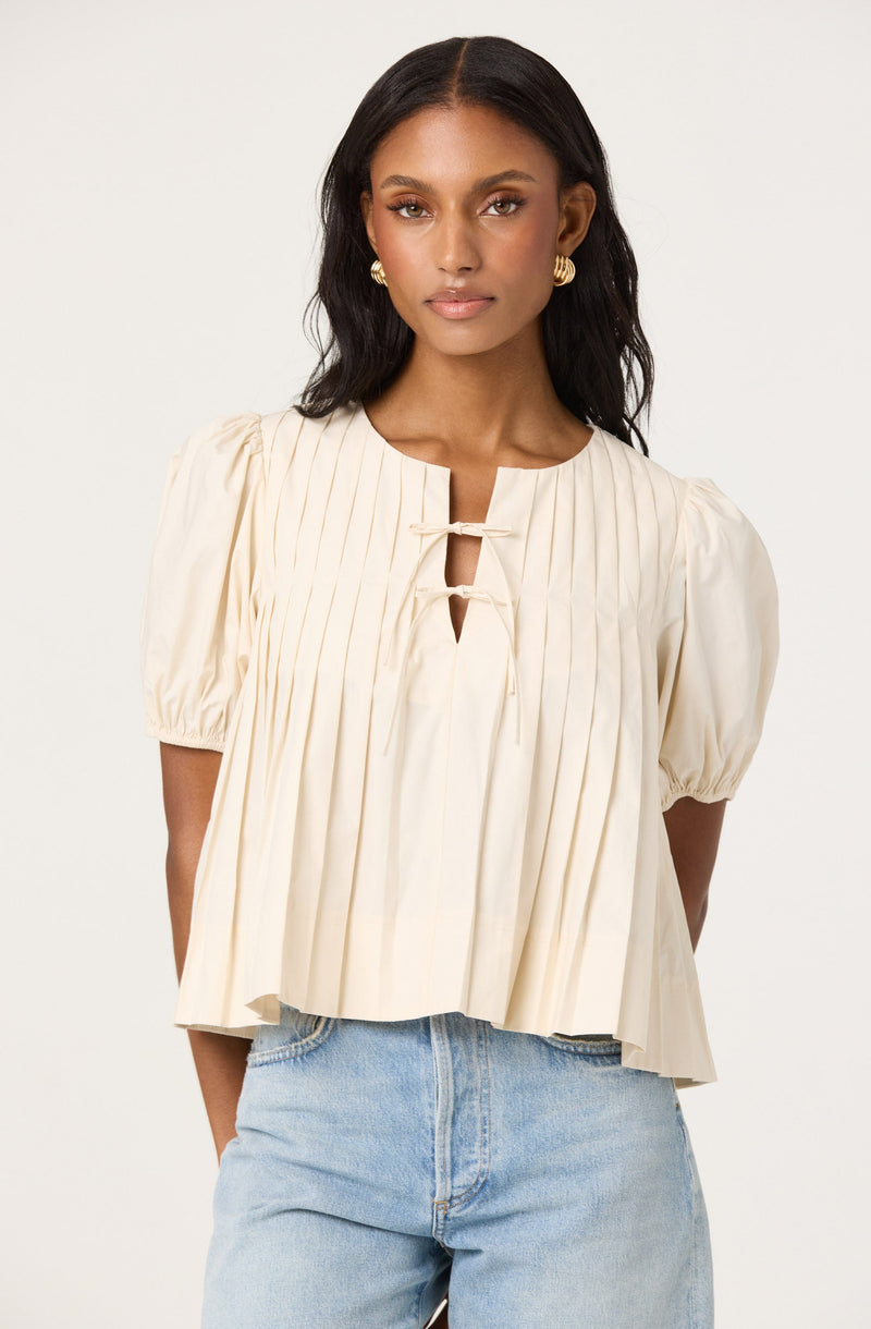 A woman is wearing a cream-colored, short-sleeved blouse with vertical pleats. The blouse features a round neckline with a small keyhole opening fastened by two fabric ties. She is also wearing light-wash denim jeans.