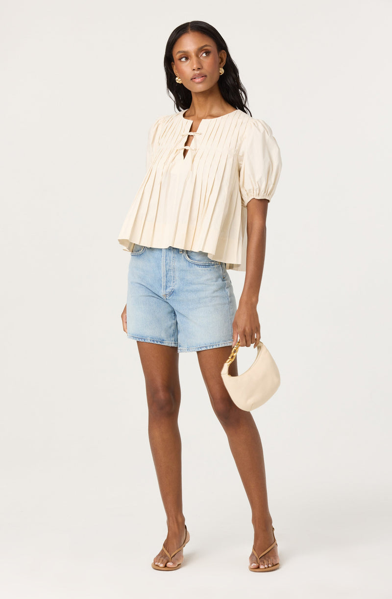 A woman is wearing a cream-colored, short-sleeved blouse with pleats and a keyhole neckline, paired with light blue denim shorts. She is holding a small, cream-colored handbag with a gold handle and is wearing clear heeled sandals.