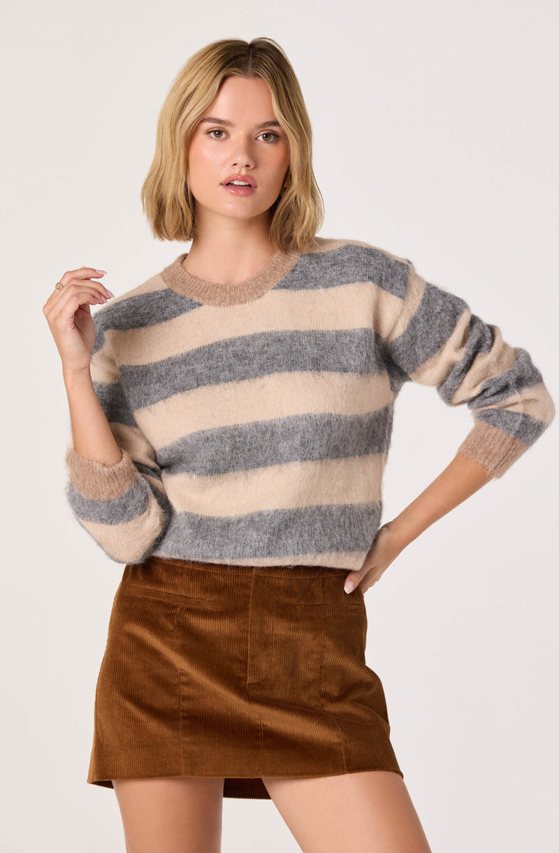 A woman is wearing a cozy, oversized knit sweater with horizontal stripes in beige and gray. The sweater has a round neckline and long sleeves. She is also wearing a high-waisted brown corduroy skirt with a front pocket.