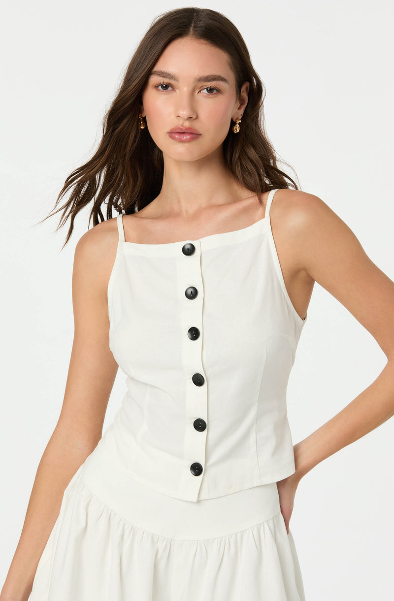 A woman is wearing a sleeveless, cream-colored top with thin spaghetti straps and a front row of black buttons. The top has a fitted silhouette and is paired with a matching cream skirt with a gathered, ruffled detail at the waist.