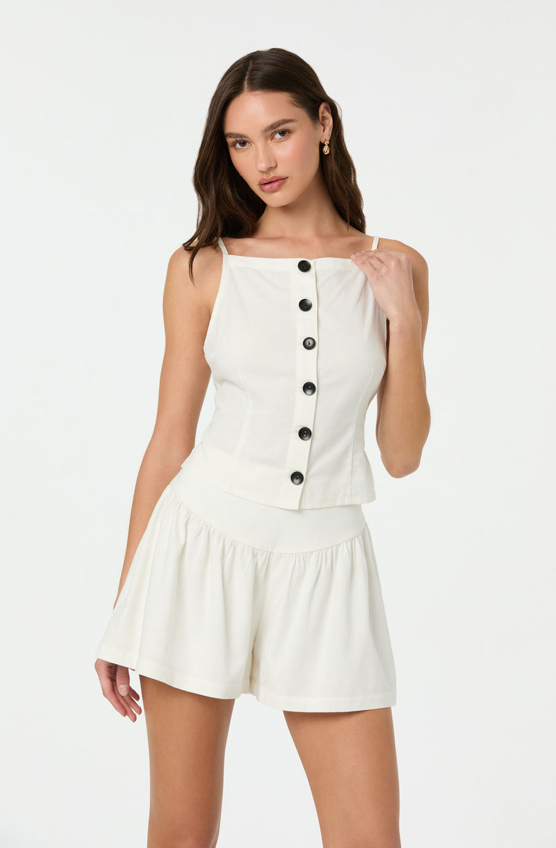A woman is wearing a sleeveless, white, button-up top with thin straps and a square neckline. The top features large black buttons running down the front. She pairs it with matching white shorts that have a gathered, elastic waistband and a loose, flowy fit.