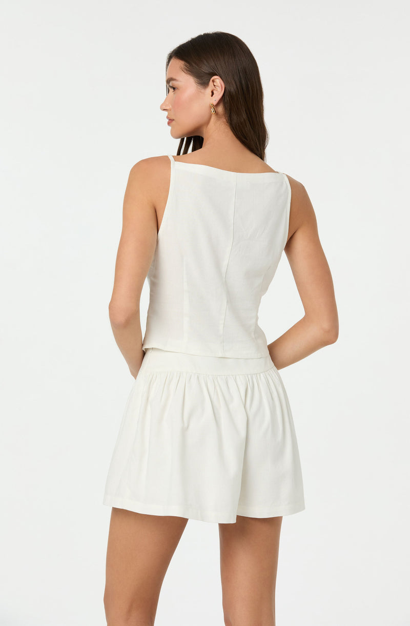 A woman is wearing a sleeveless, white top with wide shoulder straps and a boat neckline, paired with matching high-waisted, loose-fitting white shorts with a gathered elastic waistband.