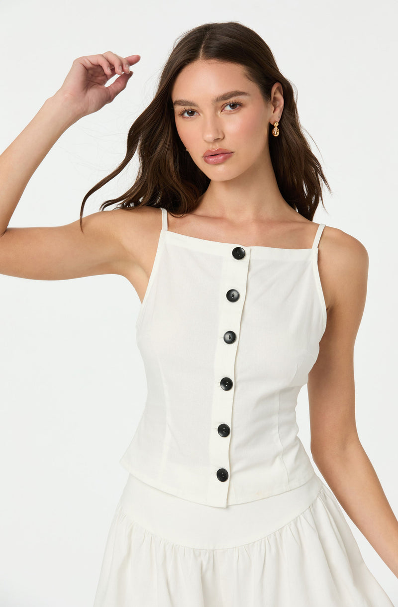 A young woman with long, wavy brown hair wearing a sleeveless white top with thin spaghetti straps. The top features a center row of large black buttons running vertically down the front. She is also wearing a pair of gold earrings.