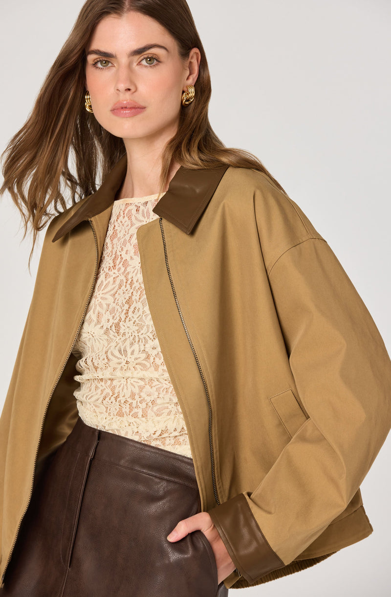 A woman is wearing a tan oversized jacket with a dark brown collar and cuffs, a cream lace top underneath, and dark brown leather pants. She is accessorized with large gold hoop earrings.
