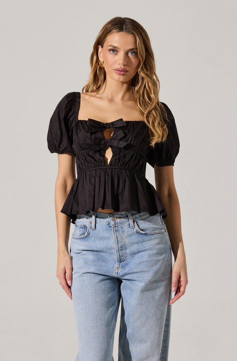 A woman is wearing a black, short-sleeved, fitted blouse with puffed sleeves and decorative bow accents on the front. The blouse has cut-out details below the bows and features a peplum-style hem. She is also wearing high-waisted, light blue jeans.