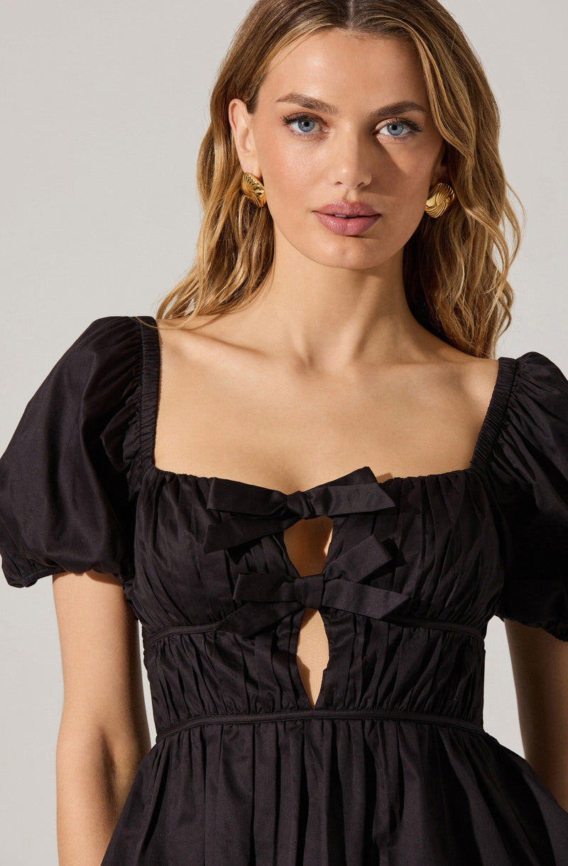 A woman is wearing a black dress with puffed short sleeves and a square neckline. The dress features ruffled detailing along the neckline and front with two bow accents and a keyhole opening below them. The fabric is gathered and cinched at the waist, creating a fitted silhouette. She has gold, textured hoop earrings and has long, wavy blonde hair.