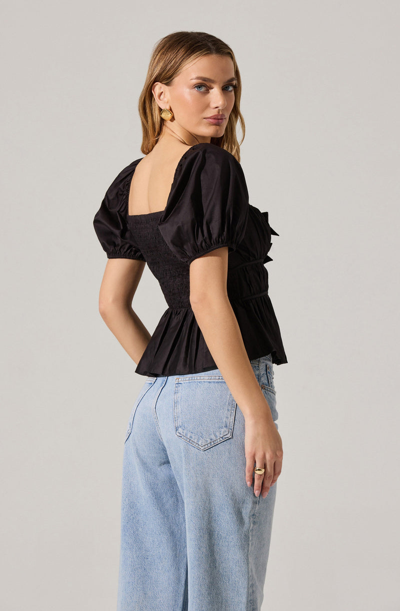 A woman is wearing a black top with short, puffed sleeves made of shiny fabric. The top features a fitted, ruched bodice with a peplum-style ruffle at the waist. She pairs it with light blue high-waisted jeans.